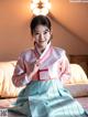 A woman in a pink and blue hanbok sitting on a bed.